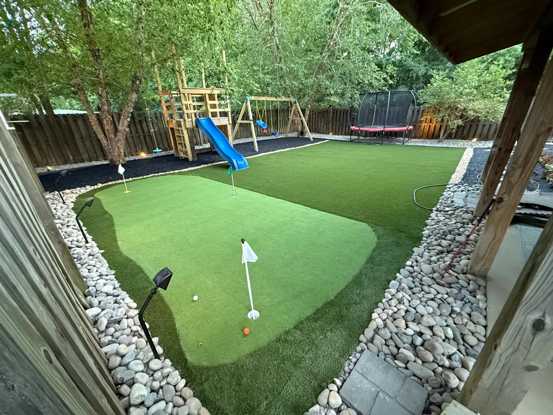 Backyard synthetic putting green edged with river rock beside a play set